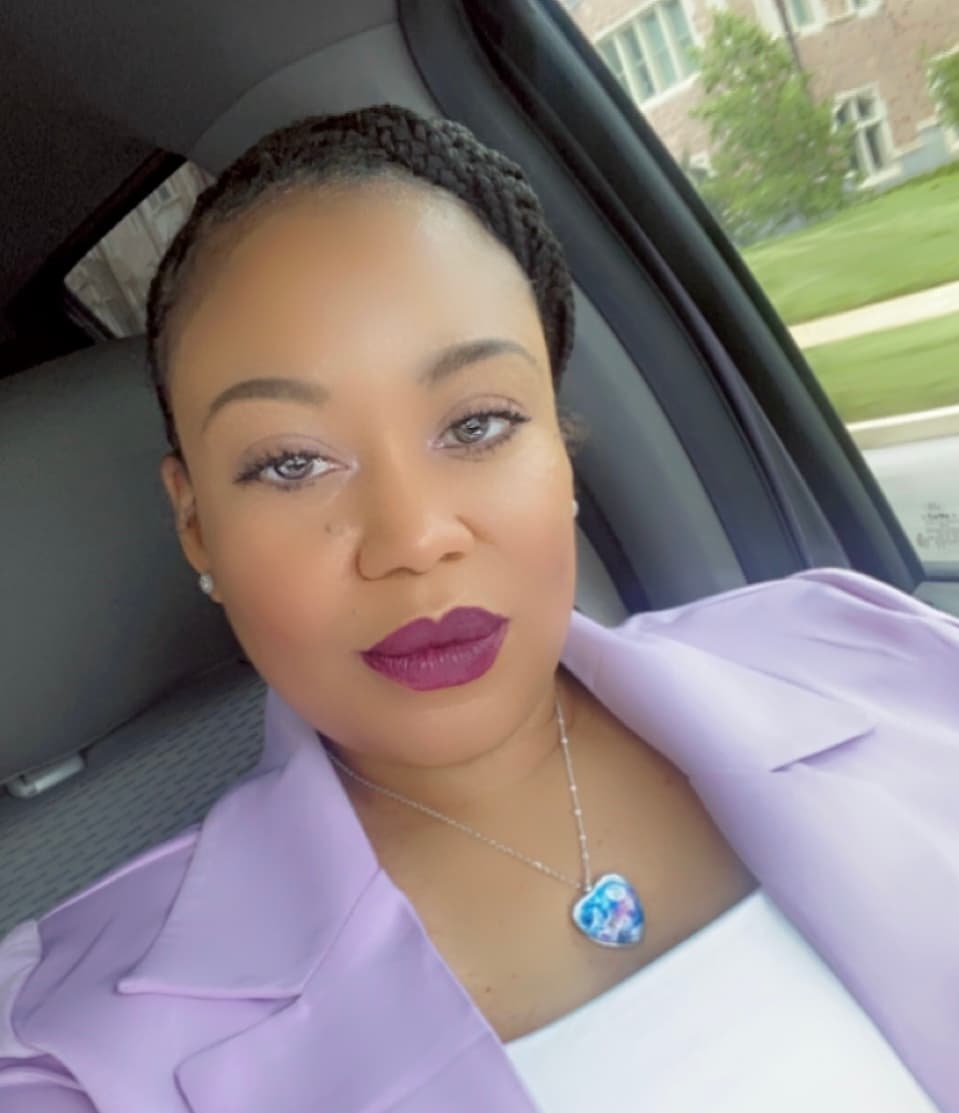 Woman with dark braids and purple lipstick wearing a lavender blazer and heart necklace.